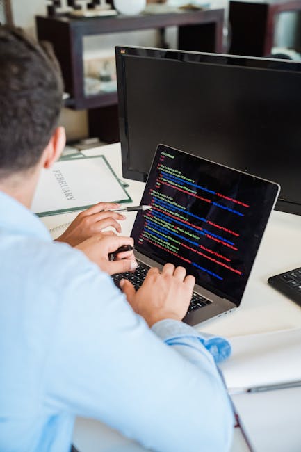 A person with short dark hair, wearing a light blue shirt, is seated at a desk and working on a laptop that displays a screen filled with colorful lines of code. The desk has multiple computer monitor